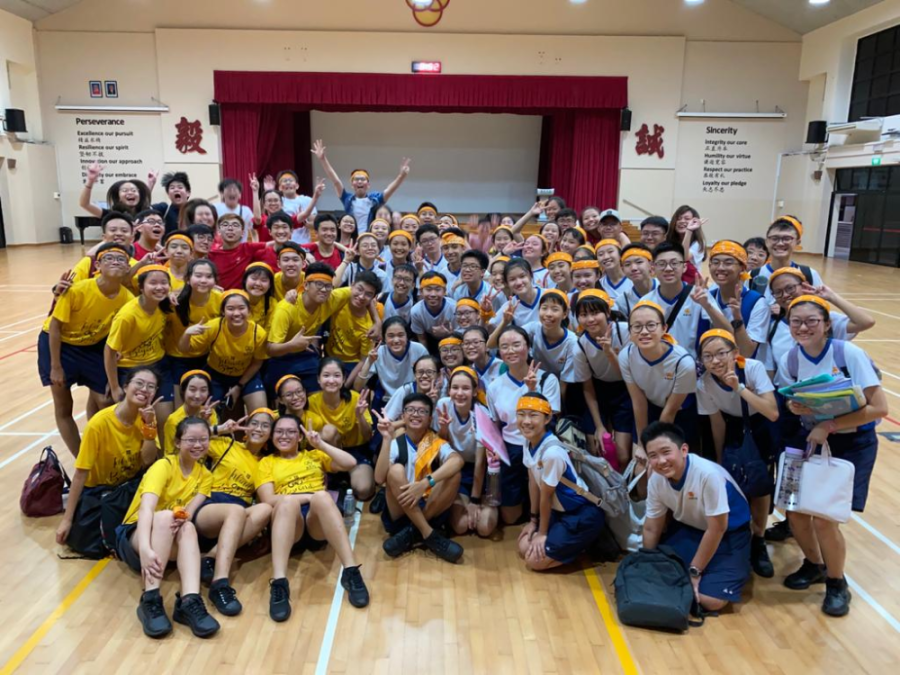 Large group of students in yellow and white uniforms with orange headbands posing cheerfully in a school hall.